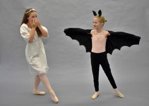 a young dancer playing a princess, dressed in white, reacts to a dancer playing a bat who poses with wings outstretched