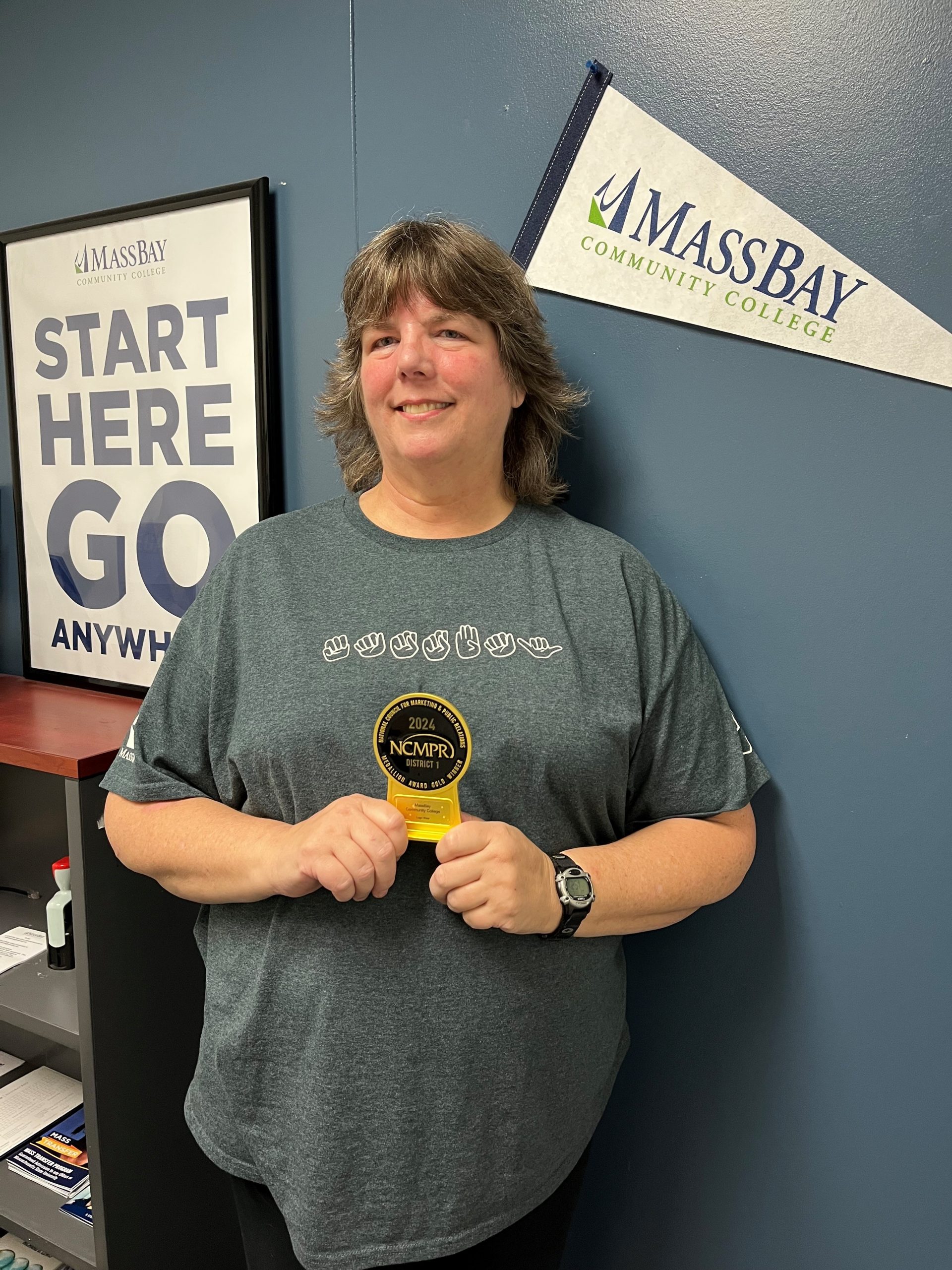 MassBay Community College Admissions staff member, Lisa Dunn, holds the 2024 National Council for Marketing and Public Relations (NCMPR) Gold Medallion award, which was in recognition of category of Logo Wear for the MassBay ASL T-Shirt. The award winning MassBay ASL T-Shirt, which Dunn is wearing, was inspired by Dunn and quickly became a favorite shirt among MassBay staff. Wellesley Hills campus, Wellesley Hills, MA, December 2024 (Photo/ MassBay Community College).