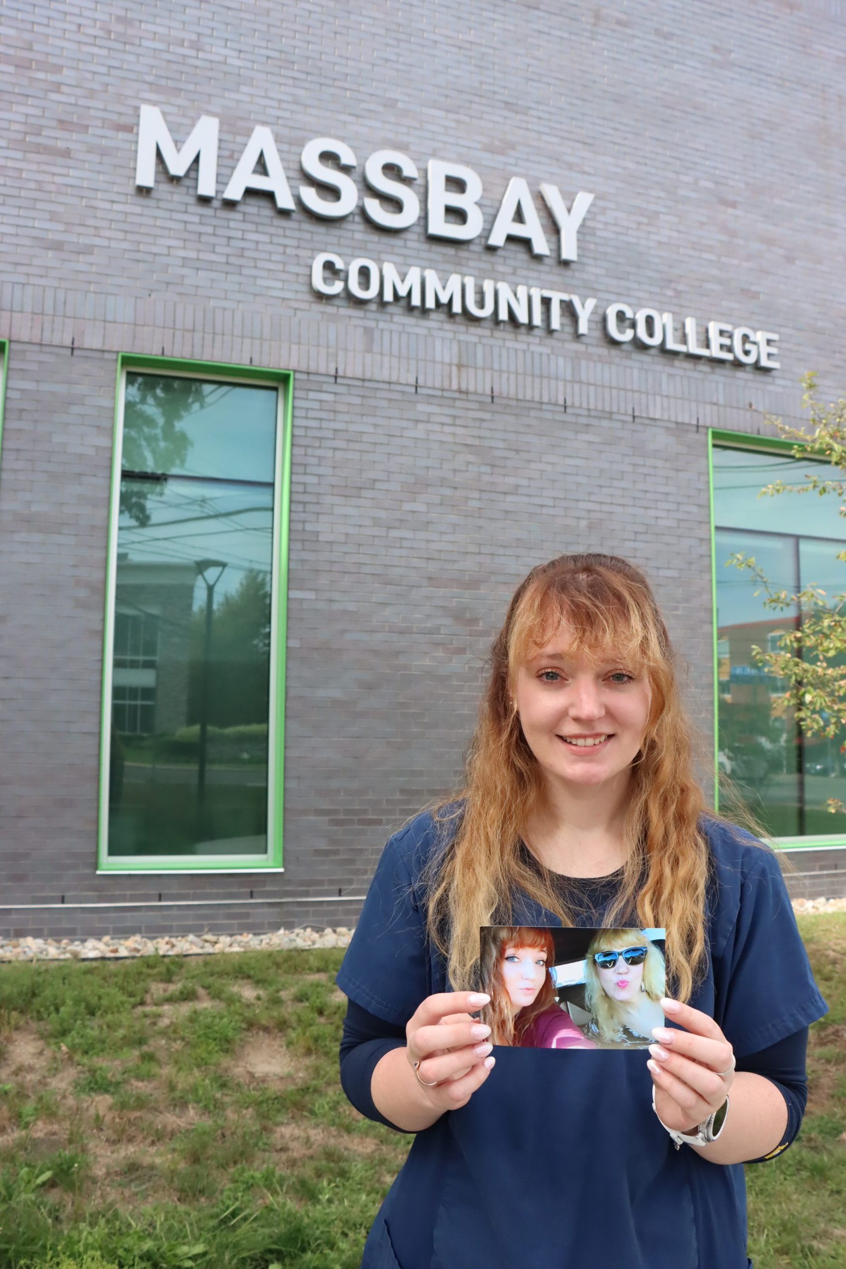 Alt text: A young woman in navy scrubs stands outside the MassBay Community College building, smiling while holding a photo of herself with her mother. The building’s large sign reading “MassBay Community College” is visible above her.