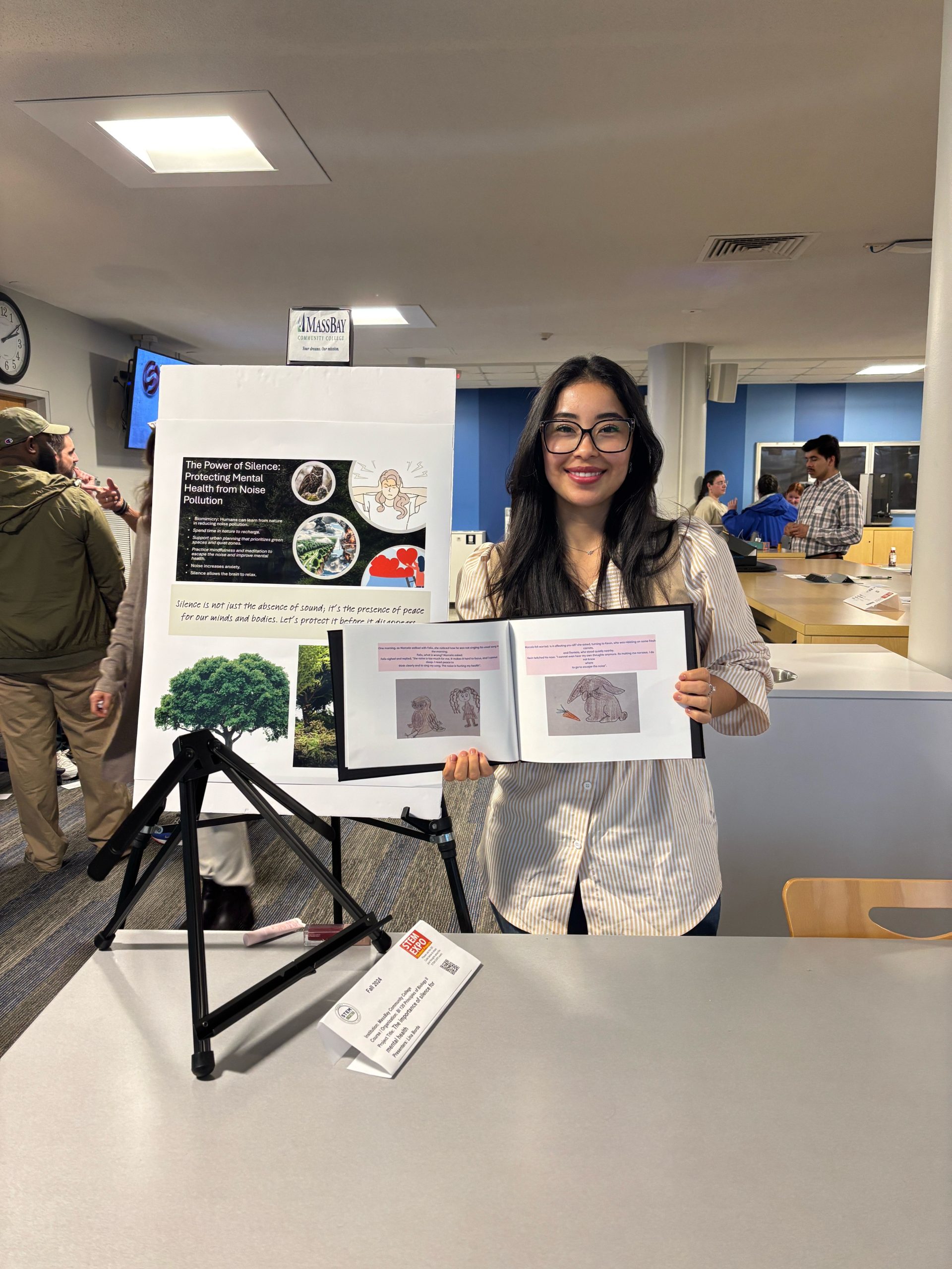 A person stands behind a table at an indoor event, holding an open illustrated book with drawings and text. Next to them is a large poster on an easel featuring images of animals, nature, and text about biomimicry and noise pollution. A folded tripod stand and a name card are on the table. In the background, several people are gathered and talking in a brightly lit room with blue walls and counters.