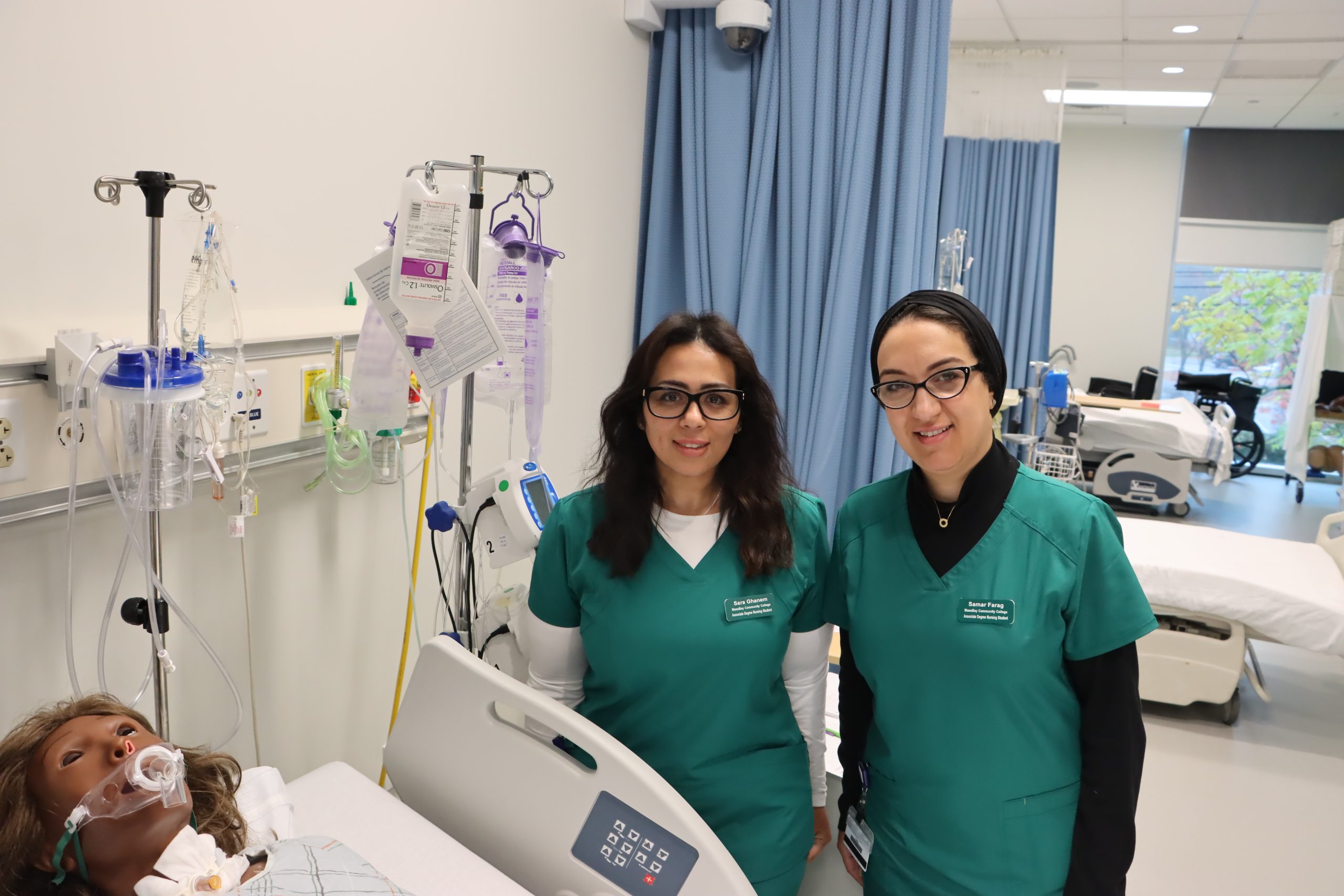Two women in green scrubs, dark hair and dark glasses, standing in a hospital room setting in the nursing lab on the Framingham campus