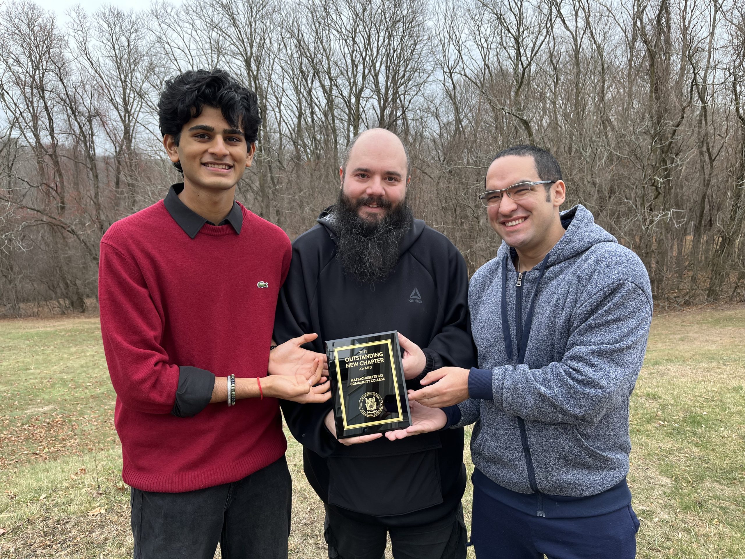 A group of three men posing for a picture, holding a plaque that says "Outstanding New Chapter", there are trees and a wooded area behind them. This photo was taken on the MassBay Wellesley Hills campus at 50 Oakland Street, Wellesley Hills.