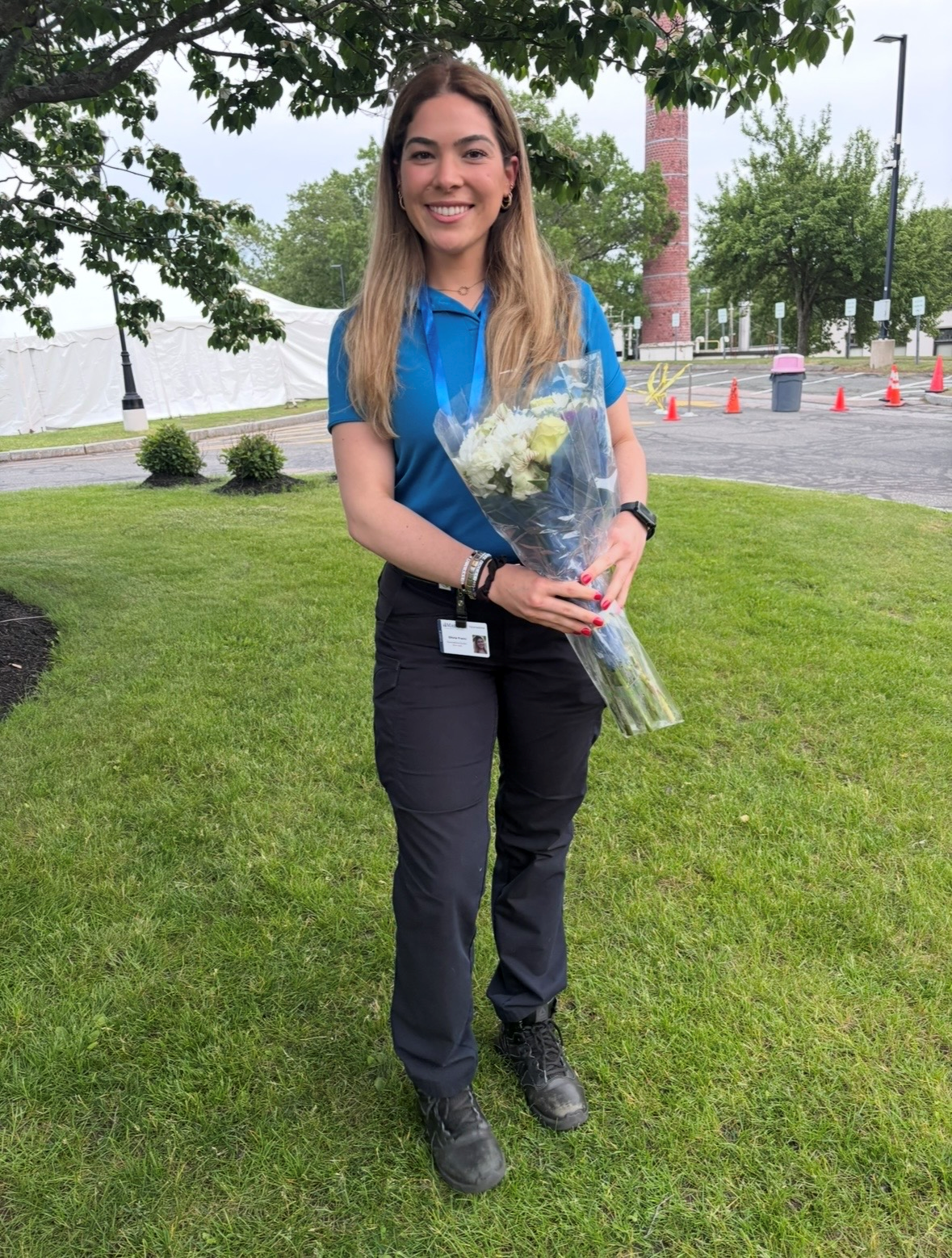 A female wearing a blue polo shirt and black pants stands on a grassy campus lawn holding a bouquet of white flowers, with trees, cones, and campus buildings visible in the background.