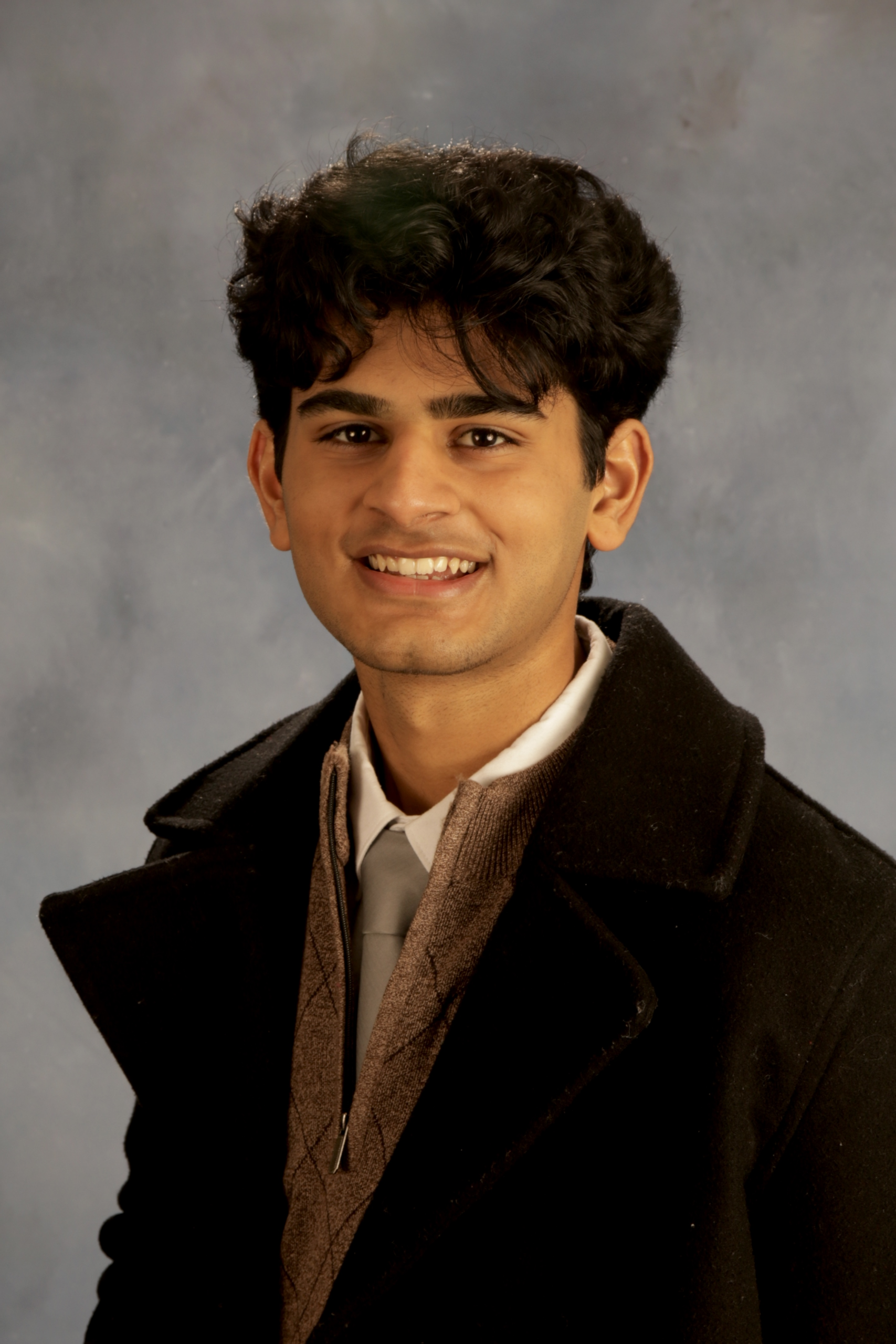 Studio-style headshot of a person wearing a dark coat layered over a textured sweater, collared shirt, and tie, posed against a neutral gray backdrop with soft, even lighting.