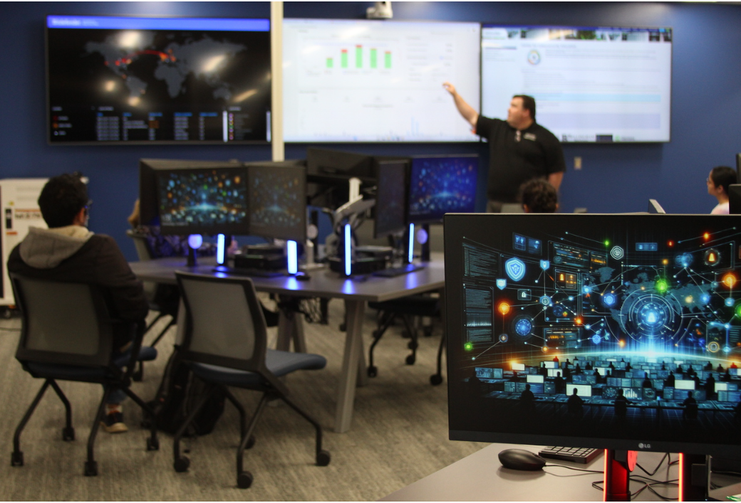 Students sit at computer workstations in a technology lab while an instructor gestures toward wall-mounted screens displaying data dashboards and charts. Multiple monitors show coding interfaces and futuristic data visualizations, highlighting a collaborative, hands-on learning environment focused on advanced computing and data analysis.