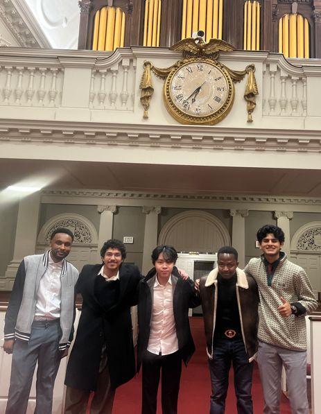 Five people stand shoulder to shoulder inside a historic hall, with a large decorative clock and organ pipes mounted high on the wall behind them. The group is positioned at the front of the room, which features classical architectural details and red carpeting.